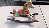 Side view of wooden decorative rocking horse showing red saddle and blue accents on cream cracked finish body, mounted on dark wood curved base.
