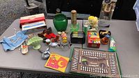 A variety of vintage items arranged on a table including glassware, figurines, board game, and packaged goods.