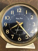 Front face of Baby Ben alarm clock showing black dial, gold numerals, and hands with brand names.