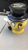 Side view of yellow ceramic jar shaped like Mr. Peanut with monocle, winking eye, and white gloved hand saluting. Black ceramic top hat lid on top.