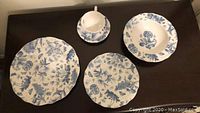 Four round ceramic plates and a cup with saucer, all with blue botanical flower patterns on white background, displayed on a brown surface.