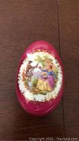 Front view of one Limoges egg-shaped porcelain box, showing detailed romantic scene artwork on lid and deep red color.