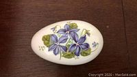 Top view of a white porcelain egg featuring detailed purple flowers and green leaves painted on the surface.