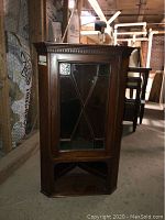 Front view of antique wooden corner wall cabinet with glass door in a storage setting.
