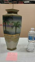 Photo showing the hexagonal shape of the vase, the hand painted palm trees and sunset scene with detailed gold geometric lattice near base, alongside a water bottle for size reference.