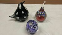 Photo showing the three art glass pieces: a black and white bird figurine, a purple swirl glass paperweight, and a colorful clear teardrop glass.