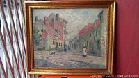 Full view of framed oil painting showing village street, buildings with red roofs, and figures in period dress.