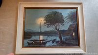 Frontal view of the framed watercolour painting showing the full riverside scene with boats, large tree, and hut lit by interior light during dusk or dawn.