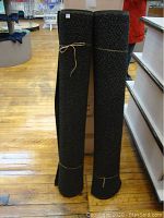 Two black heavy rubber door mats rolled up and secured with brown string, standing upright on wooden floor near counter.