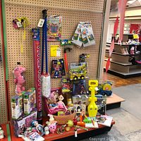 Full view of toys including dolls, jump rope, toy vehicles, packs of creativity games and plush toys displayed on shelf against pegboard.
