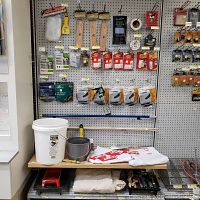 Wide view of hardware items hanging on pegboard including clips, clamps, staples, tacks, insulation tape, buckets, paint supplies, and clothing