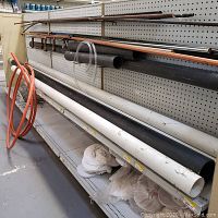 Various metal and PVC pipes arranged in a rack, including copper, galvanized steel, black and white PVC pipes, orange flexible tubing and some painter's caps on the bottom shelf.