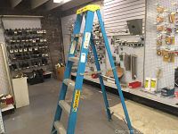 Full view of blue fiberglass step ladder standing open on workshop floor, showing metal steps, yellow top cap, and visible labels.