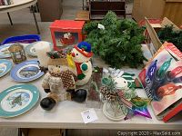 Table with 7 Avon Christmas collector plates, fabric snowman decorations, candles, lanterns, and other holiday decor