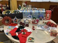 Overview of Christmas decorative items arranged on table showing stockings, mugs, wreath, ornaments, candles, mugs, carafe and glass plate