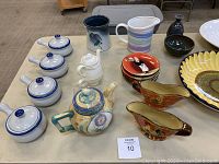 Front view of lot showing 4 lidded soup bowls with blue rim, 2 gravy boats with floral design, 2 ceramic pitchers, 2 decorative vases, 2 teapots, and 6 small dessert plates stacked.