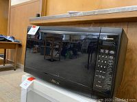 Panasonic black microwave oven with keypad visible, placed on a white box surface in a room with wooden wall paneling and tables.