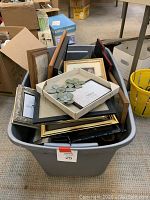 Large gray storage bin filled with multiple picture frames of different colors and designs, partially stacked and arranged haphazardly.