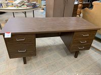Front view of a vintage wooden desk showing five drawers with metal handles and wooden round legs, placed on carpet flooring.