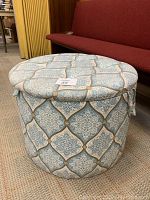 Front angle view of large round fabric ottoman with patterned upholstery in blue, white, and beige, showing general shape and fabric design.