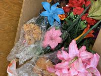 Box filled with assorted artificial flowers including blue, pink, orange, and red lilies, partially wrapped in bubble wrap.