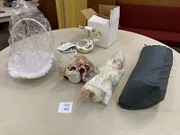 Lot items on table including two dolls, white woven basket wrapped in plastic, and dark folded item tent or fabric.