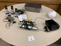 Complete lot of electronics on round table including HP printer without cord, two AT&T cordless phone sets, power strips, black CD clock radio, white spherical device, and cords.