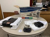 Panasonic home theater system components on a round table with white styrofoam packaging pieces. Visible items are DVD player, wireless rear speaker, cables, and accessories.
