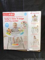 Front of box showing the Skip Hop Explore & More Baby's View 3-Stage Activity Center, features and images of the product with a baby. The box displays colorful toys attached to the activity center and mentions 25+ developmental activities.
