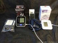 Lot overview showing three digital blood pressure monitors including EHPow AES-U181 model, white monitor, digital display monitor, cuffs, and boxes on black cloth background.