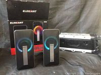 Two Elegiant SR300 PC multimedia speakers with black bodies, silver vertical central strips, showing RGB lighting effects (blue, purple) around grills, with their box in background and a rugged silver and black speaker to the side.