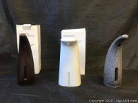 Three automatic hand soap dispensers standing upright side by side with their original boxes behind them, showing front view of each dispenser.