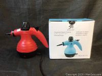 Red handheld steam cleaner next to its white and blue box with product info and branding.