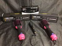 Three items on black fabric background showing two boxed One Step hot air brushes and one mini hair straightener in front, boxes labeled hot air brush and hair dryer and styler.