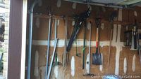 Garden tools hanging on a garage wall rack including rakes, hoes, shovels, and other metal and wooden tools showing general usage for garden maintenance.