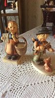 Two Hummel figurines together on a lace cloth: girl with basket and letter, boy playing violin with dog