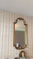 Full view of the gilt frame mirror hanging on a patterned wallpapered wall, showing its rectangular shape with curved ornamental gilt edges.