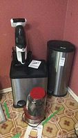 Photo showing two stainless steel trash cans (one large rectangular dual-bin and one tall cylindrical) with Shark cordless mini vacuum on charging base and instruction manual on top of rectangular trash can; Cuisinart popcorn popper with red lid and base in front on floor.