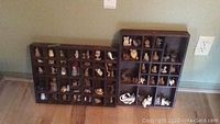Two wooden display cases filled with more than 20 small figurines, arranged in cubby compartments against a wall on the floor.