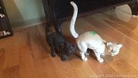 Two cast iron cat door stops, one white larger cat and one smaller black cat, positioned on a hardwood floor.