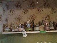 Wide view of 7 decorative music boxes displayed on a shelf with floral wallpaper background, showing varying figures including a blue-suited mouse figurine, a doll, and round boxes