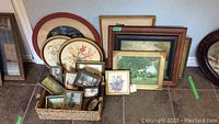 Wide shot showing multiple framed artworks of various sizes leaning against a wall including oval floral prints and landscape scenes.