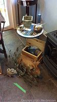 Photo showing wood pig on floor, metal peacock wall decor in background, small painted wood table holding plant pot and small decorative items, wooden drawer with glass items inside
