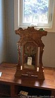 Full front view of wooden gingerbread clock on table beneath window, showing entire clock and carved detail.