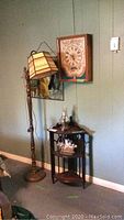Floor lamp with stained glass-style shade, wooden corner shelving unit with three curved shelves, wall mounted wooden clock with cross stitch detail, and faux stained glass wall hanging.
