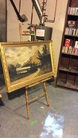 Framed mountain landscape oil painting on wooden easel in a basement setting.