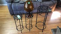 Photo showing three black metal plant stands of varying heights and one large amber-colored glass vase bowl on wooden floor.