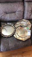 8 silver plated chargers and one anniversary platter shown stacked on a sofa with visible tarnish and reflective silver surfaces