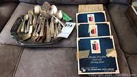 Photo of mixed silver plate flatware and silver plate platter piled on a surface with three boxed sets of silver spoons in the background.