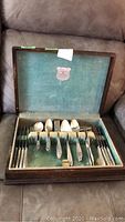 Full set of Community Plate Adam flatware arranged inside the hinged wood box with visible velvety greenish Tarnishield lining, showing variety of knives, forks, spoons, and serving pieces.